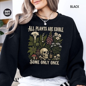 Pu&ograve; includere: Felpa nera con un'illustrazione botanica raffigurante un teschio, funghi e varie piante. Il testo recita: "Tutte le piante sono commestibili, alcune solo una volta".