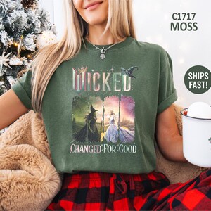 May include: Moss green t-shirt featuring the word "Wicked" in a colorful, glittery font, with an image of two witches. The phrase "Changed For Good" is printed below the image. The shirt is a comfortable, casual style.