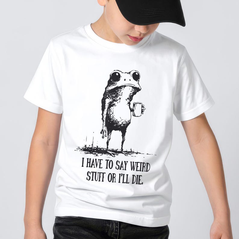 May include: White t-shirt featuring a black and white illustration of a frog holding a mug. The text "I HAVE TO SAY WEIRD STUFF OR I'LL DIE" is printed below the frog. The shirt is worn by a person.