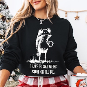 May include: Black sweatshirt with a white graphic of a frog holding a mug. The text "I HAVE TO SAY WEIRD STUFF OR I'LL DIE" is printed below the frog. The sweatshirt is part of a clothing collection.