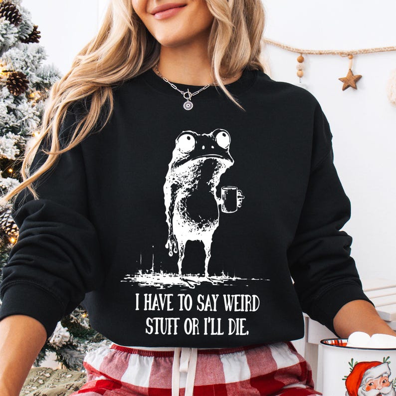May include: Black sweatshirt with a white graphic of a frog holding a mug. The text "I HAVE TO SAY WEIRD STUFF OR I'LL DIE" is printed below the frog. The sweatshirt is part of a clothing collection.