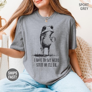May include: Sport grey t-shirt featuring a black illustration of a frog holding a mug. The frog is standing and has large eyes. The text "I HAVE TO SAY WEIRD STUFF OR I'LL DIE" is below the frog. The shirt is a casual style.