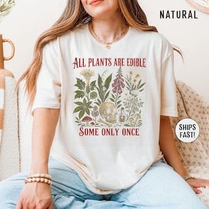Pu&ograve; includere: T-shirt color crema con un'illustrazione botanica con un teschio, funghi e varie piante. Il testo rosso dice "ALL PLANTS ARE EDIBLE SOME ONLY ONCE". La maglietta ha maniche corte.