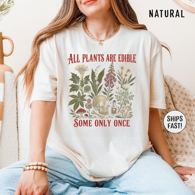 Pu&ograve; includere: T-shirt color crema con un'illustrazione botanica con un teschio, funghi e varie piante. Il testo rosso dice "ALL PLANTS ARE EDIBLE SOME ONLY ONCE". La maglietta ha maniche corte.