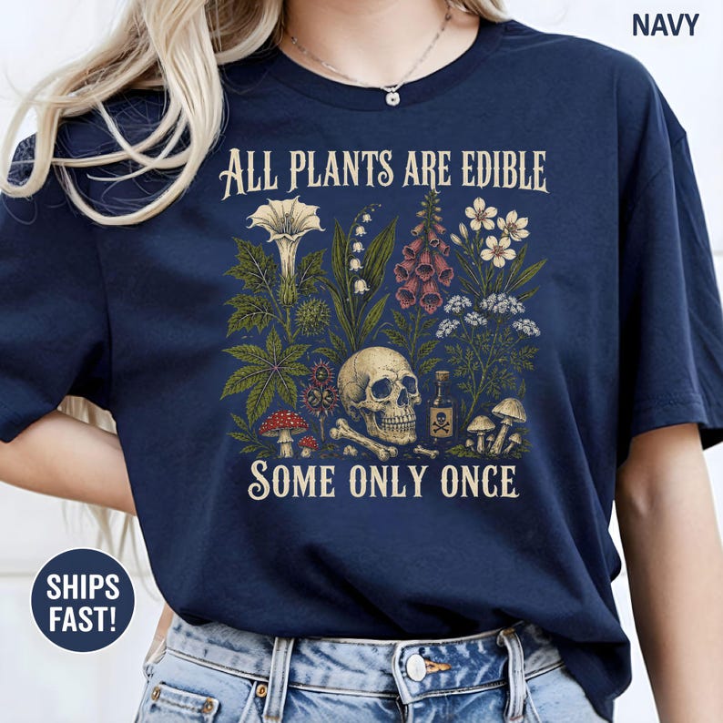 Pu&ograve; includere: T-shirt blu navy con la scritta "ALL PLANTS ARE EDIBLE SOME ONLY ONCE" e un'illustrazione botanica con un teschio, funghi e piante. Il design include una bottiglia di veleno e ossa incrociate.