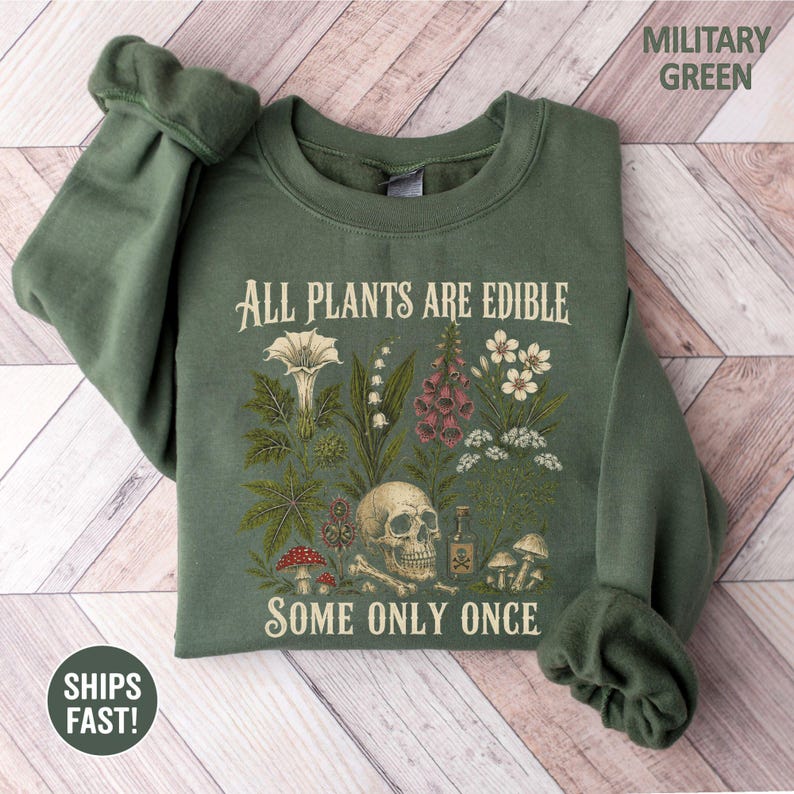 Pu&ograve; includere: Felpa verde militare con la scritta "ALL PLANTS ARE EDIBLE SOME ONLY ONCE" e illustrazioni botaniche, tra cui un teschio, funghi e fiori. La felpa &egrave; piegata su una superficie di legno.
