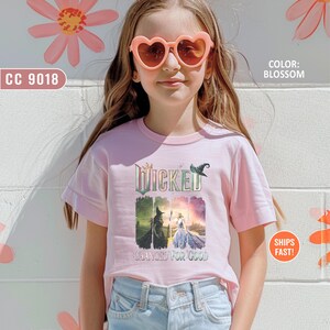 May include: A light pink t-shirt featuring the word "Wicked" with a witch hat graphic. The shirt also has the text "Changed for Good" and is worn by a person wearing heart-shaped sunglasses. The color is "Blossom".
