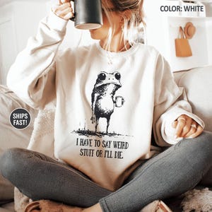 May include: White sweatshirt featuring a black and white illustration of a frog holding a mug. The text "I HAVE TO SAY WEIRD STUFF OR I'LL DIE" is below the frog. The sweatshirt is a neutral color and has long sleeves.