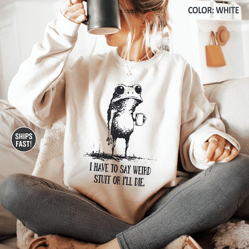 May include: White sweatshirt featuring a black and white illustration of a frog holding a mug. The text "I HAVE TO SAY WEIRD STUFF OR I'LL DIE" is below the frog. The sweatshirt is a neutral color and has long sleeves.