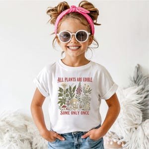 Pu&ograve; includere: Una maglietta bianca con la scritta "ALL PLANTS ARE EDIBLE SOME ONLY ONCE" in rosso. La maglietta presenta un'illustrazione botanica con fiori, funghi e un teschio. La persona indossa occhiali da sole e una fascia rosa.