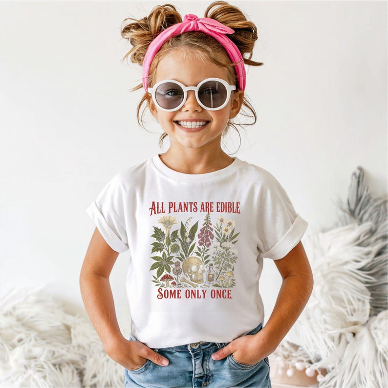 Pu&ograve; includere: Una maglietta bianca con la scritta "ALL PLANTS ARE EDIBLE SOME ONLY ONCE" in rosso. La maglietta presenta un'illustrazione botanica con fiori, funghi e un teschio. La persona indossa occhiali da sole e una fascia rosa.