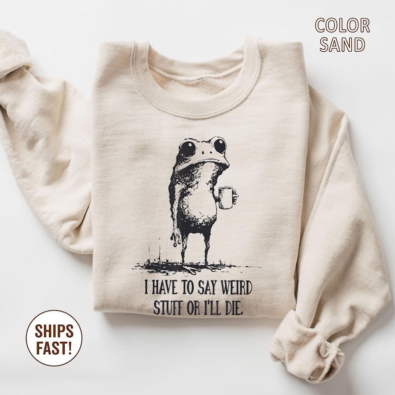 May include: Sand-colored sweatshirt with a black graphic of a frog holding a mug. The text "I HAVE TO SAY WEIRD STUFF OR I'LL DIE" is below the frog. The sweatshirt is a neutral color and has long sleeves.