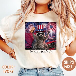 Puede incluir: Camiseta color marfil con un dise&ntilde;o de un mapache con gafas de sol y un sombrero del T&iacute;o Sam, sosteniendo un perrito caliente y una bandera estadounidense. El texto en el dise&ntilde;o dice "bad day to be a hot dog."
