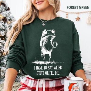 May include: Forest green sweatshirt with a white graphic of a frog holding a mug. The text "I HAVE TO SAY WEIRD STUFF OR I'LL DIE" is below the frog. The sweatshirt is a crew neck style.