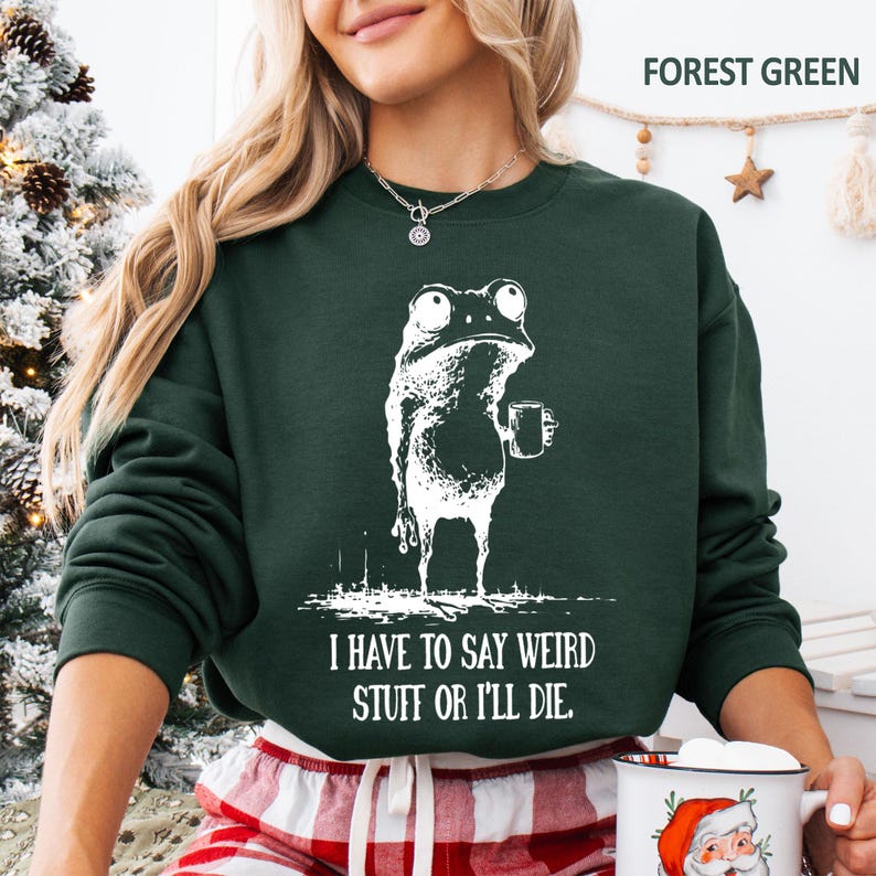 May include: Forest green sweatshirt with a white graphic of a frog holding a mug. The text "I HAVE TO SAY WEIRD STUFF OR I'LL DIE" is below the frog. The sweatshirt is a crew neck style.