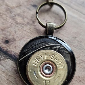 May include: A keychain with a round pendant featuring a brass shotgun shell casing with the text "HEVI-SHOT 12". The pendant has a black background with a feather design and is attached to a bronze-colored metal chain.