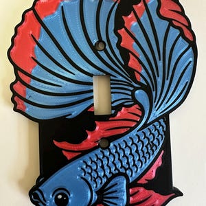 May include: A black light switch plate featuring a raised, three-dimensional design of a betta fish. The fish has a blue body and flowing fins with red edges. The plate includes a central rectangular cutout for a toggle switch and two screw holes.