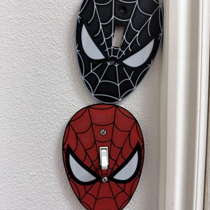 May include: Two oval light switch plates shaped like superhero masks. The top plate is black with a white web pattern and white eyes. The bottom plate is red with a black web pattern and white eyes, featuring a white toggle switch and two screws.