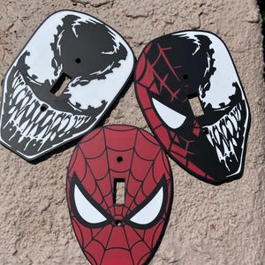 May include: Three decorative light switch plates featuring comic book character designs. One plate is black and white with a monstrous face, one is red and black with a split face design, and one is red with a black web pattern and white eyes.