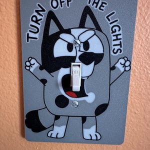May include: A gray rectangular light switch cover featuring a black and white cartoon dog illustration with the text TURN OFF THE LIGHTS printed in black. The switch toggle is white and positioned in the center of the dog's open mouth.