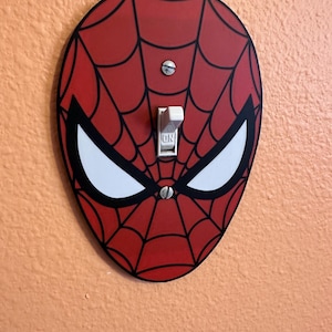 May include: An oval-shaped light switch cover plate featuring a red and black spiderweb pattern with large white eyes. The cover is secured with two silver screws and surrounds a white toggle switch with the word ON printed on the lever.