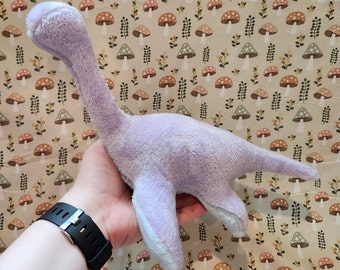 Könnte beinhalten: Ein Plüsch-Dinosaurierspielzeug in Lila mit langem Hals und Schwanz. Der Dinosaurier wird in einer Hand vor einem Hintergrund aus Stoff mit einem sich wiederholenden Muster aus kleinen Pilzen gehalten.
