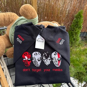 May include: Black hooded sweatshirt with embroidered skull mask designs in red, white, and black. The text "Little Devil" and "don't forget your masks" are embroidered in red. A tag reads "Koval".