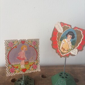 May include: Two vintage Valentine's Day card holders. One is square with a girl watering flowers, the other is heart-shaped with a woman and a feather. Both cards are mounted on green metal bases.