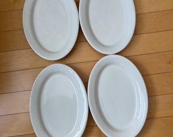 May include: Four white, oval-shaped serving platters are arranged on a wooden surface. The platters have a simple, elegant design, with a slightly raised rim. The clean, bright white color of the platters suggests a classic, versatile style.