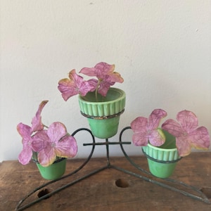 May include: A decorative metal plant stand holds three small green ceramic pots, each containing pink flowers. The stand has a heart-shaped design and sits on a wooden surface. The pots have a ribbed design.