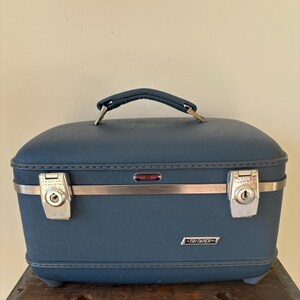 May include: A vintage blue Tri-Taper vanity case with a curved top and a handle. The case has silver-colored metal accents, including a horizontal band and two latches. The text "TRI-TAPER" is visible on the front.