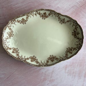 May include: Oval cream-colored serving platter with a brown floral border and trim. The platter has a scalloped edge and a slightly textured surface. The floral design is repeated around the rim.