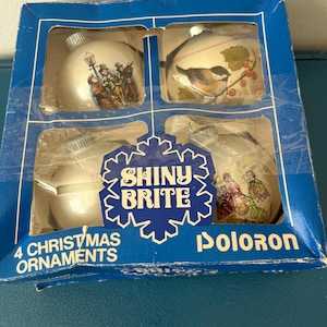 May include: A blue box with a white background containing four Christmas ornaments. The ornaments are white with different images on them. The box says "Shiny Brite" and "4 Christmas Ornaments" on it. The box also says "Doloron" on it.