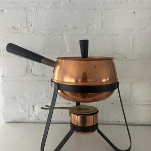 May include: Copper fondue pot with a black handle and a black metal stand. The pot has a small copper burner underneath.