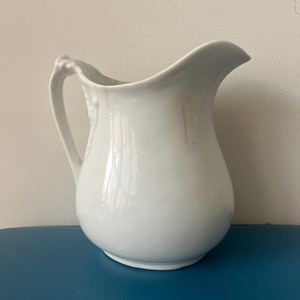 May include: A white ceramic pitcher with a curved spout and a handle. The pitcher has a rounded body and a smooth, glossy finish. The handle has a decorative design. The pitcher is sitting on a blue surface.