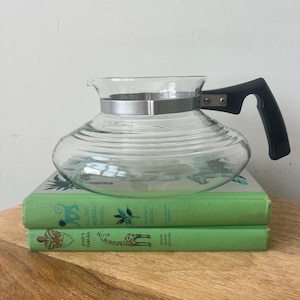 May include: A clear glass coffee pot with a black handle and a silver band around the top. The pot is sitting on top of two green books. The top book has the title "All the Mowgli Stories" and the bottom book has the title "Aesop's Fables".