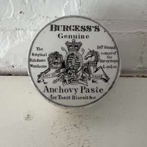 May include: A round, vintage container for Burgess's Anchovy Paste. The black and white label features a royal crest, lion, horse, and text including "Genuine" and "London." The container is for toast biscuit and is set against a white background.