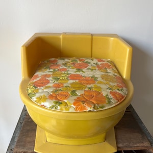 May include: A vintage yellow plastic potty chair with a floral padded seat. The seat cushion features a pattern of orange, yellow, and green flowers on a cream background. The potty chair is placed on a wooden crate.