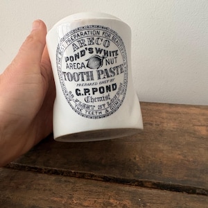 May include: A white ceramic antique tooth paste jar with a curved shape. The jar has a black circular label with text that reads "ARECO POND'S WHITE ARECA NUT TOOTH PASTE" and "C.P. POND Chemist Fleet St London".