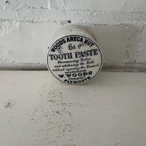 May include: A vintage, cylindrical container of "WOODS ARECA NUT TOOTH PASTE" with black text on a white background. The label includes the price "6d" and the text "For removing Tartar and whitening the Teeth without injuring the Enamel".