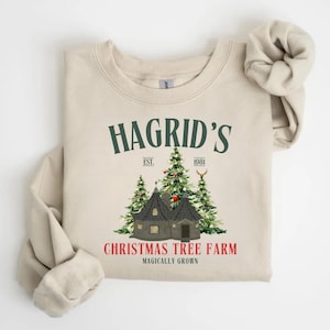 Puede incluir: Una sudadera beige con un diseño gráfico verde que dice "Hagrid's Christmas Tree Farm Magically Grown". El gráfico también incluye una pequeña casa con un árbol de Navidad en frente.