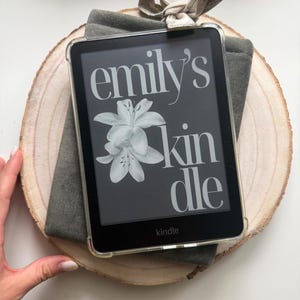 May include: A black Kindle e-reader with a clear protective case, displaying the text "emily's kindle" and a white floral design. It rests on a gray fabric sleeve and a wooden slice, tied with a beige bow.