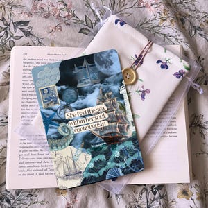May include: A book with a bookmark featuring a nautical design and the text "She had the sea within her soul, continuously." A floral pouch with a button closure is placed beside it.