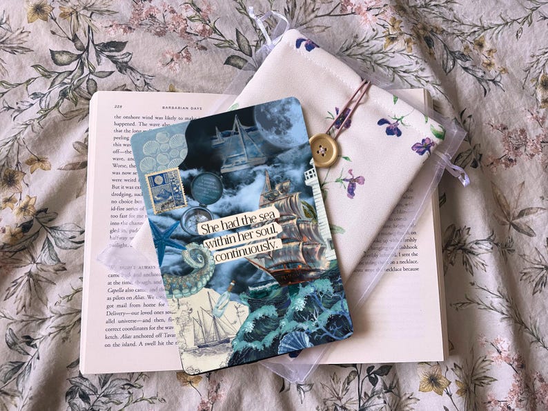 May include: A book with a bookmark featuring a nautical design and the text "She had the sea within her soul, continuously." A floral pouch with a button closure is placed beside it.