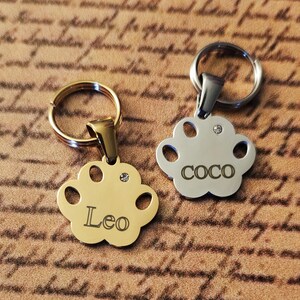 Könnte beinhalten: Zwei goldene und silberne Hundemarke in Pfotenform mit Schlüsselringen. Die goldene Marke trägt die Aufschrift "Leo" und die silberne Marke die Aufschrift "Coco".
