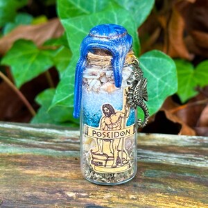 May include: A small glass bottle filled with seashells, sand, and herbs, sealed with a blue wax top. A seahorse charm and a label with the word "POSEIDON" and an illustration of a man with a trident are attached to the bottle.