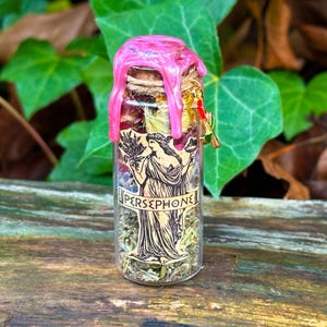 May include: A small glass bottle filled with dried herbs and flowers, sealed with a pink wax top. The bottle features a label with the word "PERSEPHONE" and an illustration of a woman. A small charm hangs from the bottle's neck.