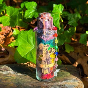 May include: A decorative glass bottle filled with layers of colorful materials, including dried flowers and crystals. The bottle features an angel illustration and the word "IRIS". A beaded charm with rainbow-colored beads and a silver butterfly hangs from the bottle.