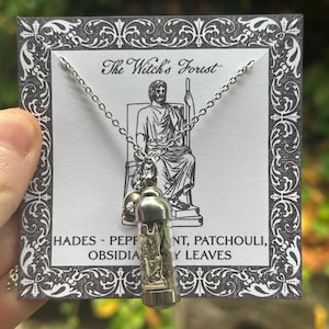 May include: A silver necklace with a small glass vial pendant. The vial contains a small statue and dried leaves. The card behind the necklace reads "The Witch's Forest" and lists ingredients. The background is blurred green.