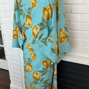 May include: A light blue kimono featuring a repeating pattern of golden yellow tulip-like flowers and muted green leaves. The garment has wide sleeves, a high collar, and a straight-cut silhouette that extends to the floor, displayed on a mannequin.